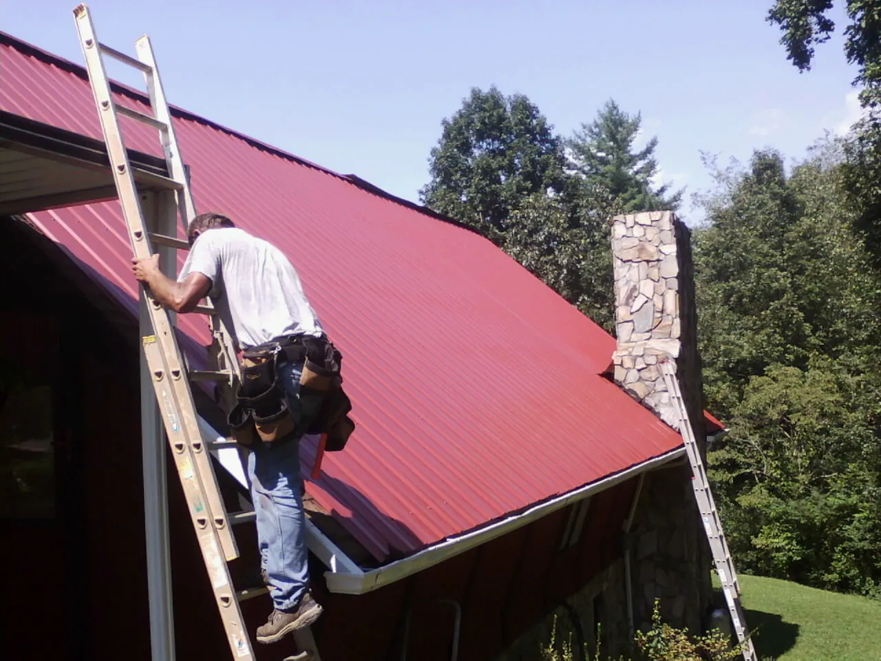 Roofer on a ladder beside a red metal roof and stone chimney — Soffit & Fascia Repair by Roofing Brothers in East Chicago
